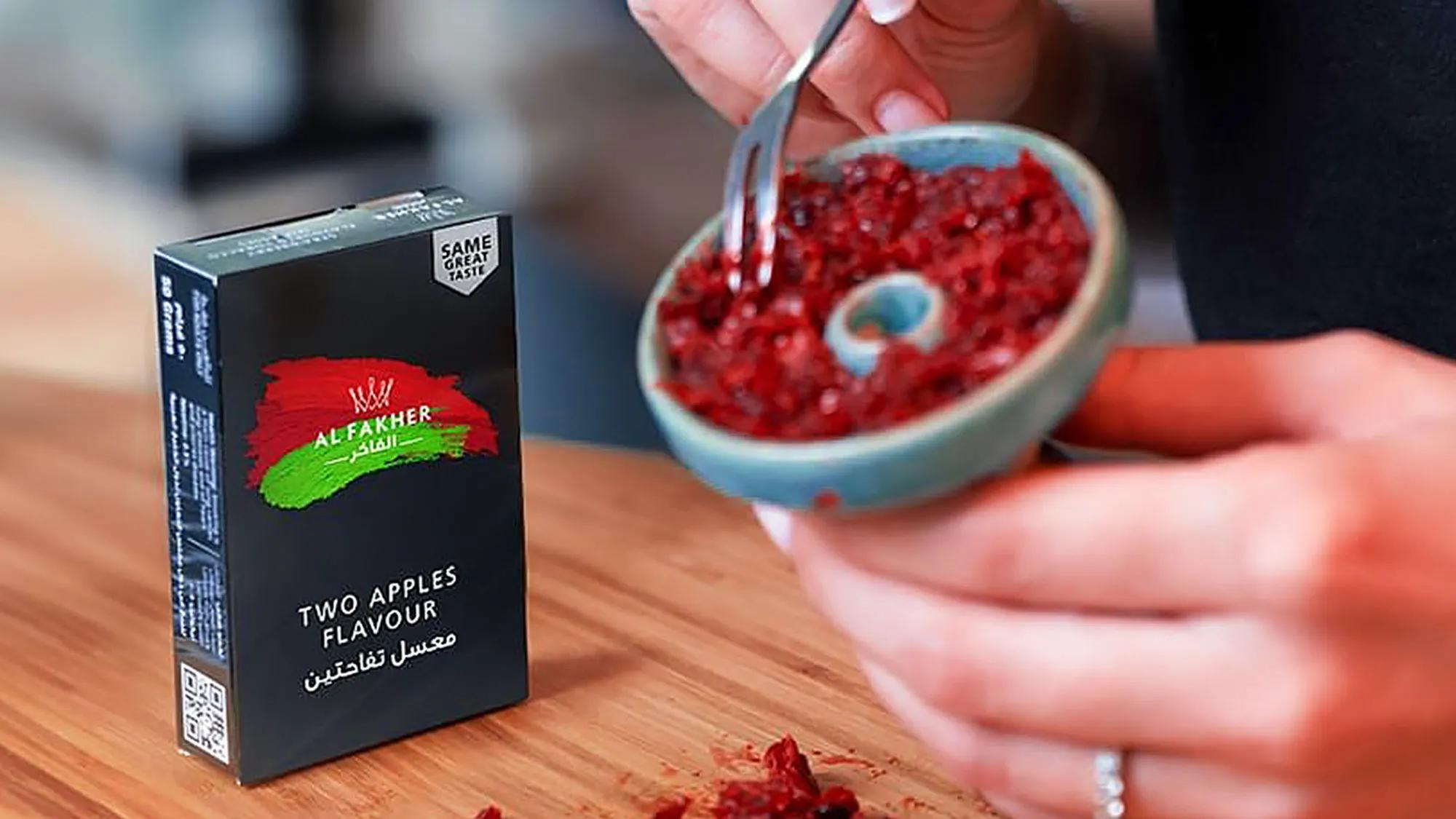 A person is preparing shisha tobacco from a box labeled Alfakher Two Apples Flavour, using a fork to scoop it from a small bowl.