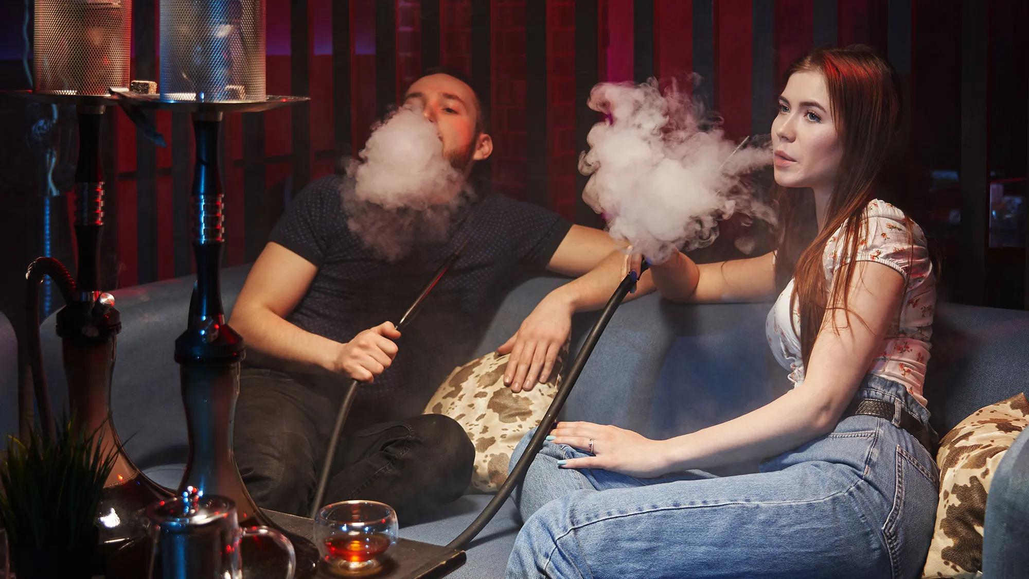 A man and a woman sit on a couch, smoking hookah. Smoke billows around them, with a cozy atmosphere and dim lighting.